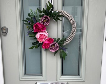 May include: A pink and purple floral wreath with greenery hangs on a light gray door. The wreath is made of woven twigs and features pink and purple peonies.