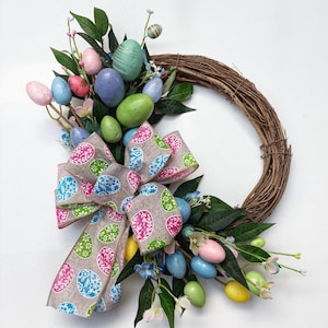 May include: A brown twig wreath decorated with pastel coloured Easter eggs, green leaves, and a pink and green patterned bow.
