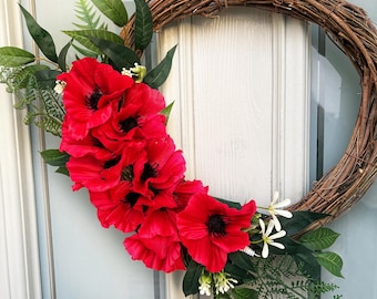 May include: A decorative wreath featuring a circular grapevine base adorned with vibrant red poppies, green leaves, and small white flowers. The wreath is hanging on a white door with a glass panel.