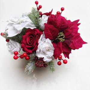 Könnte beinhalten: Ein festliches Blumenarrangement mit roten Weihnachtssternen, Rosen und Beeren, akzentuiert mit weißen Blumen und Tannenzweigen. Das Arrangement ist eine Mischung aus Texturen und Farben, perfekt für die Weihnachtsdekoration.