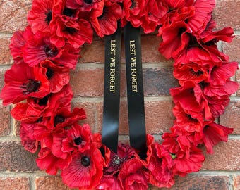 Peut inclure: Une couronne circulaire composée de nombreuses fleurs de coquelicot rouge vif avec des centres sombres. Deux rubans noirs pendent verticalement du centre de la couronne, chacun portant le texte doré LEST WE FORGET en lettres capitales.