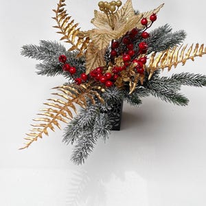 May include: A festive floral arrangement in a black square container. The arrangement features gold poinsettias and fern leaves, frosted evergreen branches, and bright red berries. A decorative piece for the holiday season.