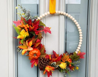 May include: A fall-themed wreath with a wooden bead ring, adorned with colorful autumn leaves, a small pumpkin, pinecone, and yellow and orange flowers. The wreath hangs on a white door with a yellow ribbon.