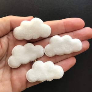 May include: Four small, white, cloud-shaped appliques with a soft, fuzzy, plush texture. Each piece features a rounded, multi-lobed silhouette resembling a stylized cumulus cloud. The items are white in color and have a consistent, uniform appearance.