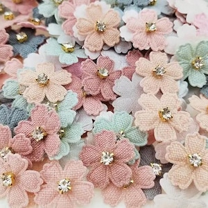 May include: A collection of pastel-coloured fabric flower embellishments with a rhinestone centre. The flowers are in shades of pink, blue, and green.