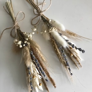 Pode incluir: Dois pequenos buquês de flores secas amarrados com barbante. Os buquês são feitos de trigo, lavanda, flores brancas fofas e outras ervas secas.