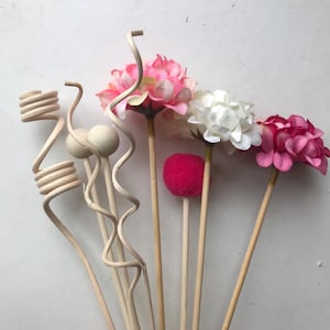 Pode incluir: Uma coleção de palitos difusores decorativos e flores artificiais. O sortido inclui palitos de madeira com desenhos em espiral e bola, juntamente com cabeças de flores rosa, brancas e fúcsia e um pompom vermelho.