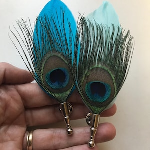 May include: Two peacock feather lapel pins. One feather is bright turquoise, the other is light blue. Both have a dark brown and teal eye pattern. The pins have silver-coloured metal bases.