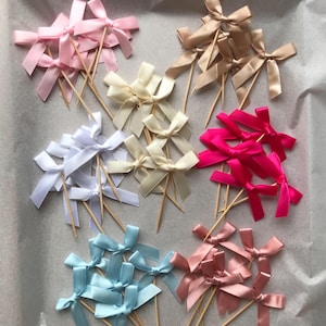 May include: Assortment of satin ribbon bows attached to wooden skewers. The bows are in various colors, including pink, white, cream, blue, and red.