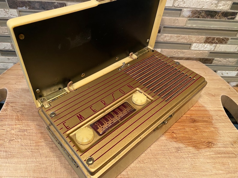 K&ouml;nnte beinhalten: Vintage Emerson-Radio mit cremefarbenem Geh&auml;use und goldfarbener Frontplatte. Das Radio hat eine rechteckige Form mit einem Klappdeckel. Die Frontplatte zeigt das Emerson-Logo und ein Zifferblatt. Das Radio ist ge&ouml;ffnet und zeigt den Lautsprecher und die internen Komponenten.