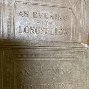 May include: Close-up of two vintage book covers. The top cover reads "AN EVENING WITH LONGFELLOW" in embossed lettering. The bottom cover reads "AN EVENING WITH HAWTHORNE". Both covers are a muted, neutral color.