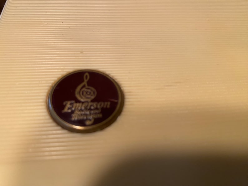 K&ouml;nnte beinhalten: Cremefarbene, geriffelte Oberfl&auml;che mit einem kreisf&ouml;rmigen Emblem. Das Emblem ist weinrot mit goldenem Rand und tr&auml;gt den Namen "Emerson" in einer stilisierten Schrift, zusammen mit zus&auml;tzlichem Text. Der Hintergrund ist hellcremefarben.