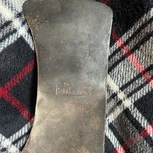 May include: A vintage axe head with a dark, weathered appearance. The metal has a worn, textured surface with areas of rust. The word "COLLINS" is stamped into the metal. The axe head is set against a plaid background.
