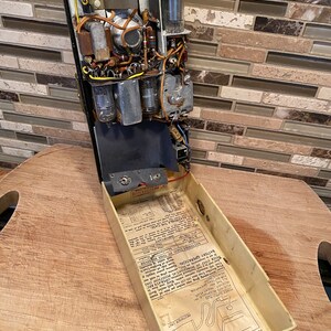 K&ouml;nnte beinhalten: Vintage Emerson-Radio mit offenem Geh&auml;use, das interne Komponenten wie Vakuumr&ouml;hren, Kondensatoren und Verkabelung zeigt. Das Geh&auml;use des Radios ist schwarz, mit einem cremefarbenen Innenraum, der Betriebsanweisungen enth&auml;lt. Ein schwarzes Netzkabel ist sichtbar.