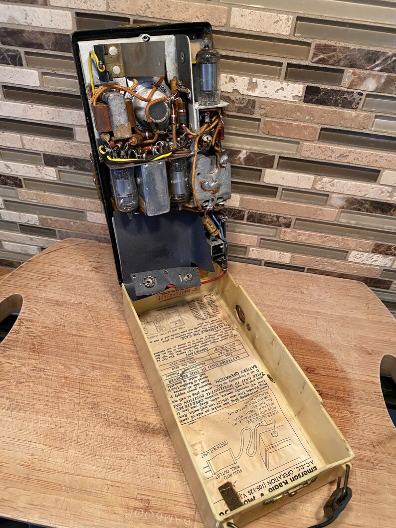 K&ouml;nnte beinhalten: Vintage Emerson-Radio mit offenem Geh&auml;use, das interne Komponenten wie Vakuumr&ouml;hren, Kondensatoren und Verkabelung zeigt. Das Geh&auml;use des Radios ist schwarz, mit einem cremefarbenen Innenraum, der Betriebsanweisungen enth&auml;lt. Ein schwarzes Netzkabel ist sichtbar.