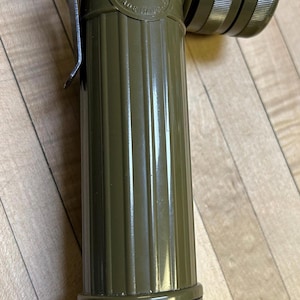 May include: Olive drab flashlight with a right-angle head. The flashlight has a ribbed body and a textured end. The text "FULTON MADE IN USA" is visible on the head. A metal clip is attached to the side.
