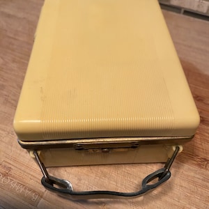 K&ouml;nnte beinhalten: Ein rechteckiger, cremefarbener Vintage-Koffer mit gerippter Textur. Er verf&uuml;gt &uuml;ber einen Metallverschluss, einen kleinen kreisf&ouml;rmigen Akzent auf der Oberseite und einen Metallgriff. Das Geh&auml;use scheint aus einem haltbaren Kunststoffmaterial zu bestehen.