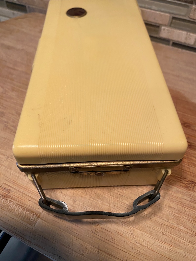K&ouml;nnte beinhalten: Ein rechteckiger, cremefarbener Vintage-Koffer mit gerippter Textur. Er verf&uuml;gt &uuml;ber einen Metallverschluss, einen kleinen kreisf&ouml;rmigen Akzent auf der Oberseite und einen Metallgriff. Das Geh&auml;use scheint aus einem haltbaren Kunststoffmaterial zu bestehen.