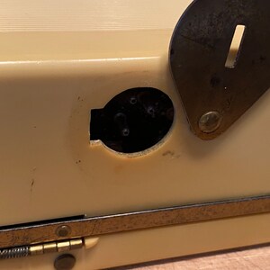 K&ouml;nnte beinhalten: Nahaufnahme eines cremefarbenen Vintage-Objekts mit einer dunklen, oval geformten &Ouml;ffnung und einem angelenkten, dunklen Metallverschluss. Der Verschluss hat einen rechteckigen Schlitz. Das Objekt hat eine messingfarbene Metallverkleidung.