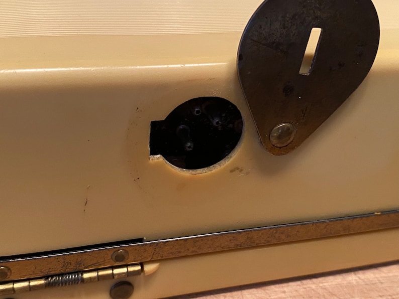 K&ouml;nnte beinhalten: Nahaufnahme eines cremefarbenen Vintage-Objekts mit einer dunklen, oval geformten &Ouml;ffnung und einem angelenkten, dunklen Metallverschluss. Der Verschluss hat einen rechteckigen Schlitz. Das Objekt hat eine messingfarbene Metallverkleidung.