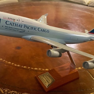 May include: A model airplane of a Cathay Pacific Cargo Boeing 747, displayed on a wooden stand. The plane is white with blue and green accents, and the text "CATHAY PACIFIC CARGO" is visible on the fuselage. A small plaque is at the base.