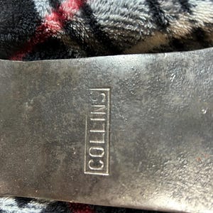 May include: Close-up of a vintage axe head with the word "COLLINS" stamped in a rectangular frame. The axe head is made of dark metal and shows signs of wear. The background is a textured fabric.