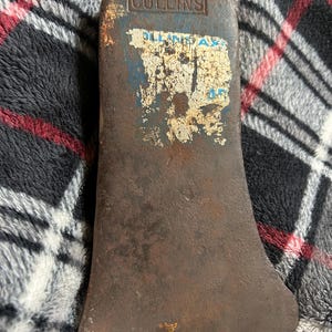 May include: A vintage axe head with a weathered, rusty appearance. The axe head has a curved blade and a rectangular top with the word "COLLINS" printed on it. A faded blue and white sticker is partially visible.