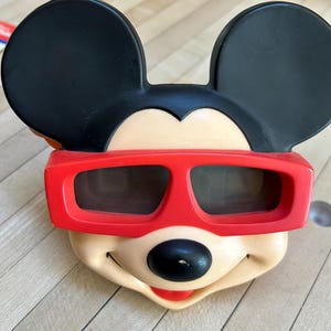 May include: A vintage Mickey Mouse face with black ears, a cream-colored face, and a red mouth. The character is wearing red 3D glasses with dark lenses. The image is a close-up of the character's face.