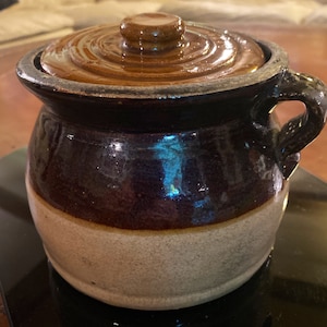 May include: A small, antique stoneware crock with a lid. The crock is a deep brown color with a lighter tan base and a dark handle. The lid is a matching brown color. The crock is likely used for food storage.