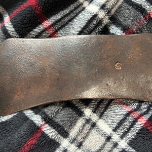 May include: A vintage, weathered axe head with a curved blade. The metal has a dark, rusty brown patina, with a small circular marking near the center. The axe head is set against a plaid background.