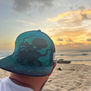 May include: A person wearing a blue and green mesh trucker hat with a black octopus graphic. The hat has a black brim and a white logo on the side. The person is looking out at a beach scene with a sunset in the background.