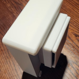 May include: A white, rectangular, 3D-printed object with a stepped design. The object has a smooth, matte finish and appears to be a component or part of a larger structure. The item is sitting on a wooden surface.