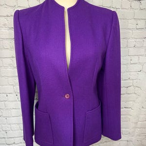 May include: A vibrant purple blazer featuring a mandarin collar, long sleeves, and a single button closure at the front. The jacket includes two patch pockets positioned at the lower front, structured shoulders, and a clean, solid-colored design.