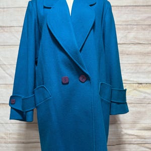 May include: A teal double-breasted coat with purple buttons and cuff accents. The coat features a classic lapel collar and two front pockets. The sleeves have a cuff detail with a single purple button. The coat is displayed on a mannequin.