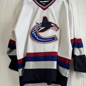 May include: White hockey jersey with a V-neck collar and the Vancouver Canucks logo on the chest. The jersey features blue, red, and silver stripes on the sleeves and bottom hem. The collar has a red and silver trim.