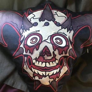 May include: A black throw pillow featuring a graphic illustration of a stylized, cracked skull with large, wide eyes, dark horns, and red accents dripping from the eye sockets and mouth. The design is centered on the fabric with a bold red outline.