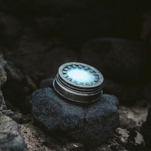 May include: A silver metal container with a blue light shining through the top. The container is sitting on a dark gray rock.