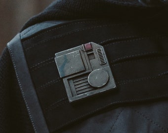 May include: A silver-colored metal pin with a rectangular shape and a circular vent. The pin has a red button and the word "MAGNETIC" is printed in yellow below the pin.