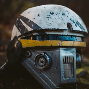 Peut inclure: Un casque blanc et gris avec des accents jaunes. Le casque a un aspect vieilli avec des gouttelettes d'eau visibles. Le casque a une visière et une petite fenêtre ronde sur le côté.