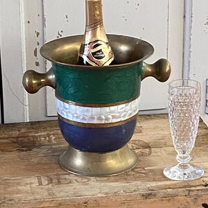 Op de afbeelding: Een champagne-emmer van messing met groene, witte en blauwe geverfde strepen. De emmer heeft twee handvatten en er zit een fles champagne in.