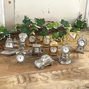 Könnte beinhalten: Eine Sammlung von elf Miniatur-Quarzuhren in verschiedenen Formen, darunter ein silbernes Schiff, ein goldenes Fahrrad, ein silbernes Auto, eine Kutsche und mehrere klassische Tischuhren. Jede Uhr hat ein weißes Zifferblatt mit schwarzen Zahlen.