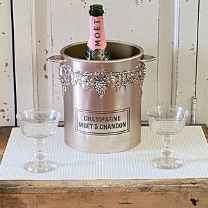 May include: A silver champagne bucket with a bottle of Moët & Chandon champagne inside, flanked by two clear champagne coupe glasses. The bucket has a decorative grape vine design and the words "CHAMPAGNE MOËT & CHANDON" are printed on the front.