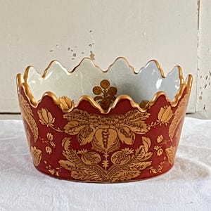 Könnte beinhalten: Dekorative Keramikschale mit gewelltem Rand. Die Schale hat einen tiefroten Hintergrund mit einem goldenen Blumenmuster. Der Innenraum ist hellblau mit goldenen Verzierungen.