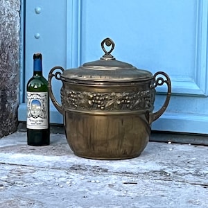 Può includere: Un piatto da portata vintage con coperchio in ottone con dettagli di grappoli d'uva e manici laterali. Una bottiglia di vino con un'etichetta blu è accanto al piatto. Lo sfondo è una porta azzurra.