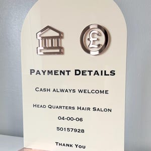 May include: An arched cream-coloured acrylic sign with rose gold icons of a bank building and a pound sterling symbol. The text reads: PAYMENT DETAILS, CASH ALWAYS WELCOME, HEAD QUARTERS HAIR SALON, 04-00-06, 50157928, THANK YOU. It has a rose gold base.