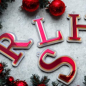 May include: Festive Christmas decorations spelling "RLHS". The red, reflective letters have gold edging and are set against a backdrop of red baubles and evergreen foliage. A seasonal, decorative item.
