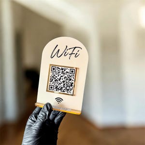 May include: A cream-coloured WiFi sign with a gold-framed QR code and the word "WiFi" in black script. The sign also features a WiFi symbol at the bottom. The sign is held by a gloved hand.