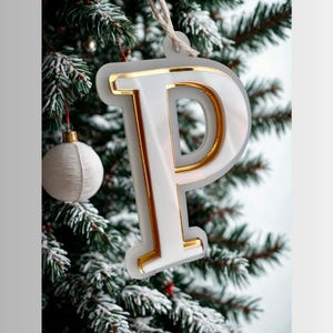 May include: A white and gold letter "P" Christmas decoration suspended from a snowy fir branch. The decoration has a gold trim and a pearlescent centre. A white bauble hangs nearby.