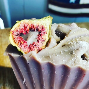 May include: A lavender-coloured soap bar with a dried fig half on top. The soap has a fluted design and is sprinkled with white crystals. The fig's interior is a vibrant red.