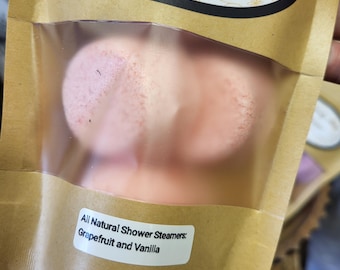 May include: A brown paper package with a clear window containing pink shower steamers. The package has a label that reads "All Natural Shower Steamers: Grapefruit and Vanilla." The top of the package has the text "Beauty Bee Soaps, Handmade & All Natural."